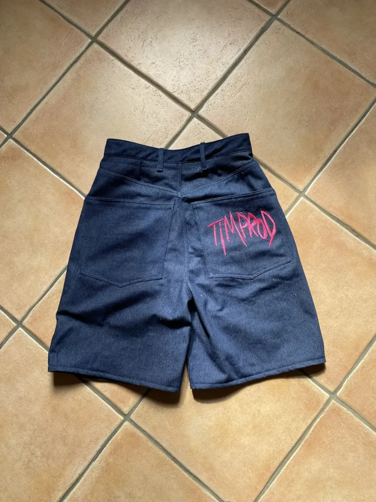 Short Timprod en denim bleu posé à plat, vu de dos, logo brodé en rose sur la poche arrière